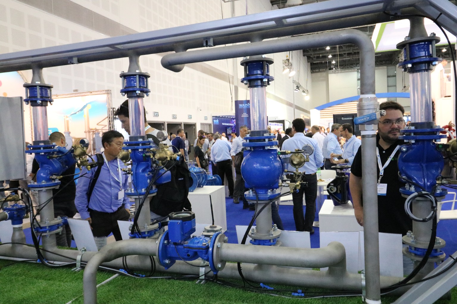 Blue flanged valves and transparent vertical pipe sections connected by grey pipework on a busy water technology exhibition floor